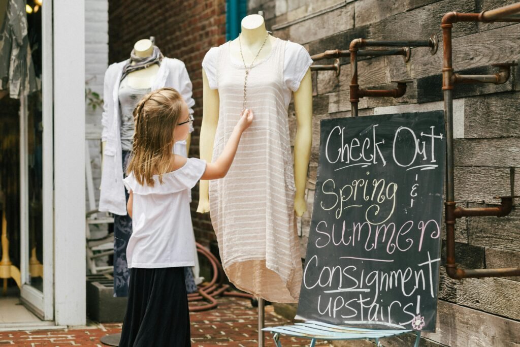 A young girl touches a mannequin in front of a clothing store with a consignment sale sign.